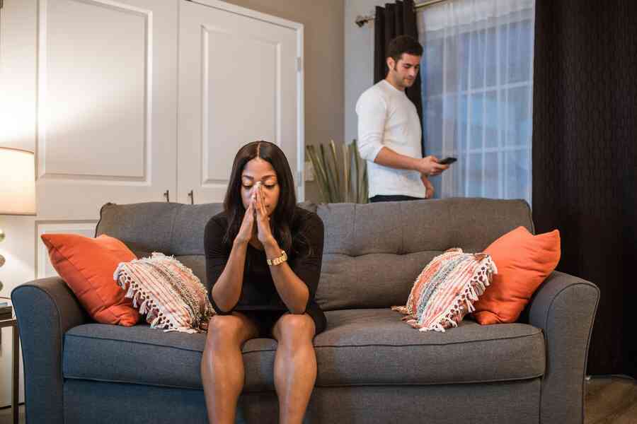 A woman looks distressed by a man standing behind her as she sits on a sofa.